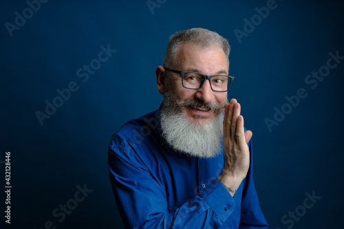 Bearded middle age man dressed blue shirt holds hand near mouth, secret talk concept, blue background