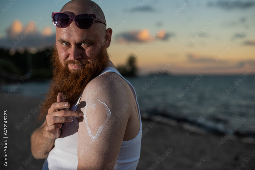 Bearded man applying sunscreen on body while standing on the beach.