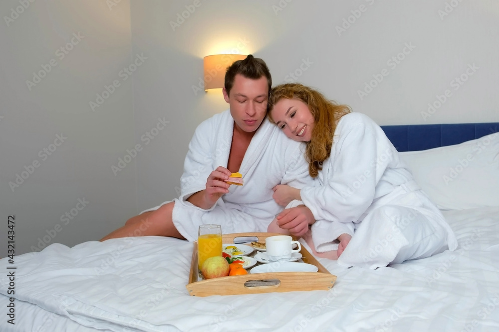 Family on vacations resting in their room. Happy young couple in bathrobes having a breakfast together sitting in bed in hotel. Man and woman are eating and drinking tea. He is feeding her.