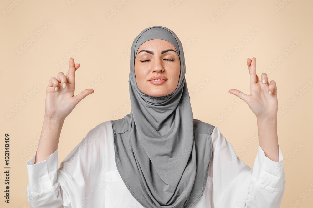 Hope concept, young arabian middle eastern muslim islamic pretty woman in grey hijab crossing fingers for good luck, hopefully expecting a miracle isolated over beige background. Superstition gesture