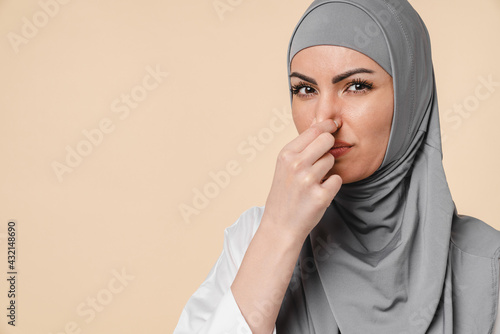 Young Middle eastern islamic muslim arabian woman wearing grey hijab isolated over beige background smelling stinky and disgusting, intolerable smell, holding breath with fingers on nose. Bad smell