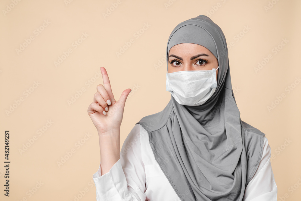 Young Middle eastern islamic muslim arabian woman in protective face mask against Covid 19 coronavirus and grey hijab pointing at copy space isolated over beige background. Young Middle eastern islamic muslim arabian woman in protective face mask against Covid 19 coronavirus and grey hijab pointing at copy space isolated over beige background.