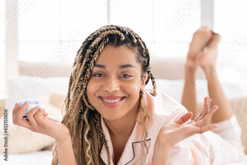 Happy relaxed young african woman lying on the bed applying anti-age cream moisturizer on her face, body care, skin care, rejuvenation concept photo