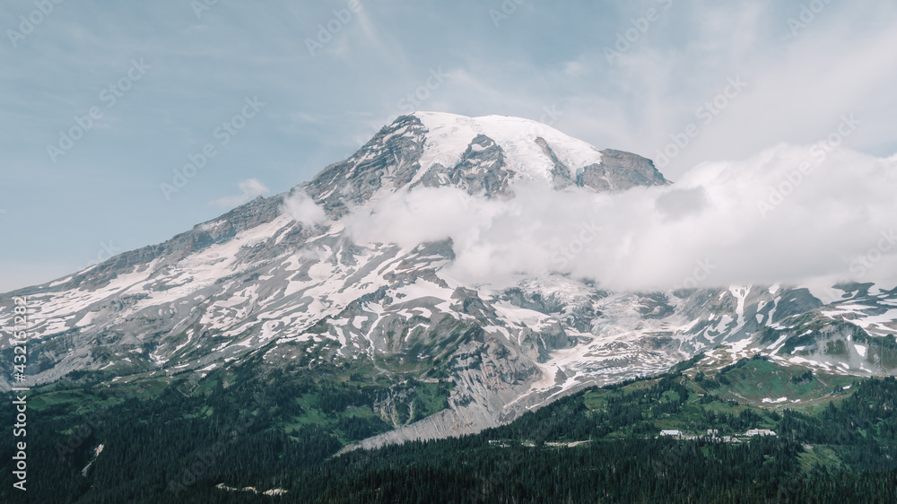 Panorama of white fluffy clouds lie just below the snowy peaks of Mount Rainier with a forest of surrounding pine trees in Mount Rainier National Park, Washington, USA.