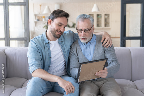 Caring loving adult son hugging embracing his old elderly senior father while he is showing his family photograph photo at home, telling stories of his youth. Happy father`s day!