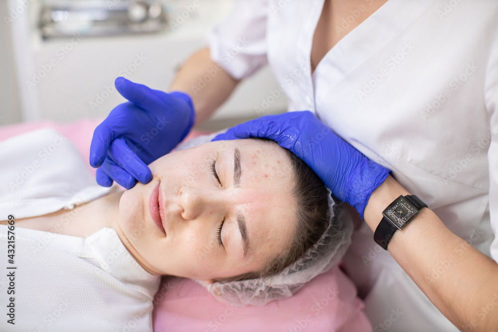 Young pleasant woman lying on the couch at dermatology center and smiling, while hands of doctor cosmetologist in rubber gloves touching her cheeks while applying lotion or serum Young pleasant woman lying on the couch at dermatology center and smiling, while hands of doctor cosmetologist in rubber gloves touching her cheeks while applying lotion or serum