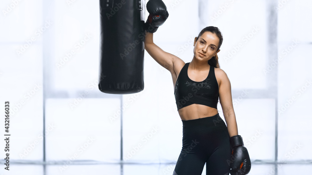 exhausted sportswoman in boxing gloves and sportswear standing near punching bag in gym.
