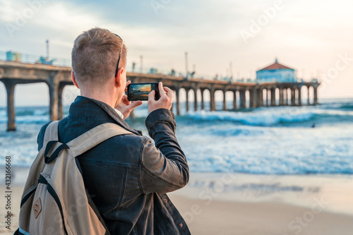 A young man with a backpack takes photos of the pier with his mobile phone on the beach in Los Angeles with a bright sunset