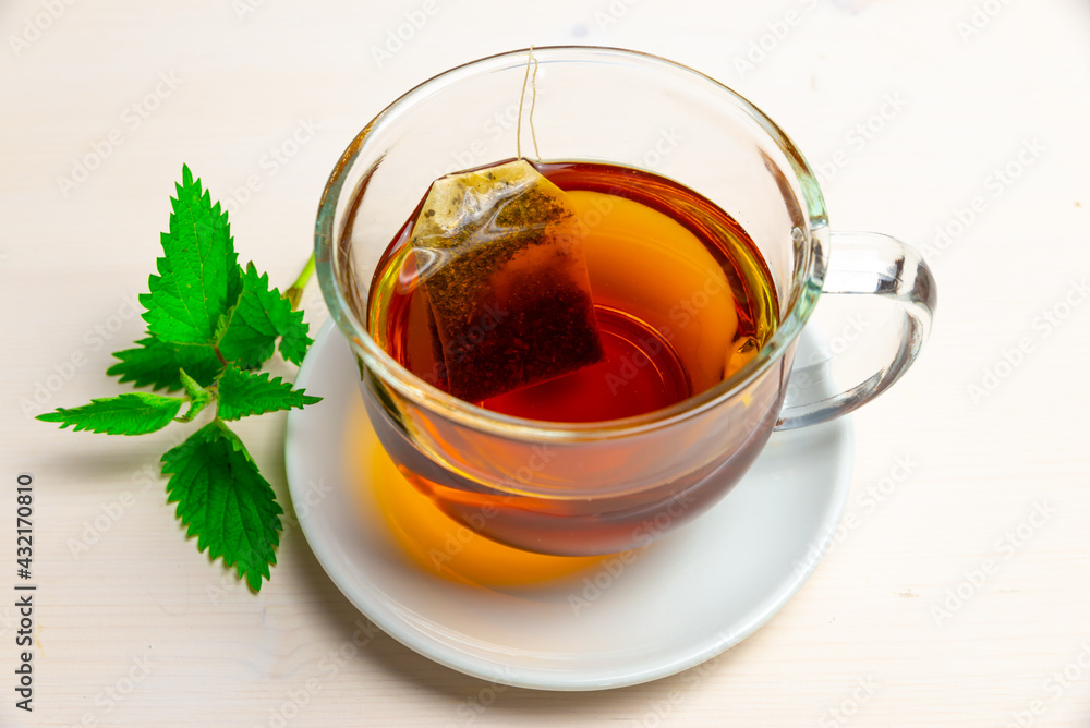Nettle herbal tea in a transparent cup, with a sachet immersed in water, and a white saucer with nettle leaves next to it. Healing infusion of nettle. 
