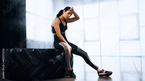 tired young sportswoman wiping sweat and sitting on tire while resting in sports center.