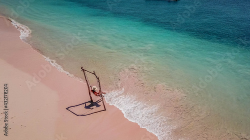 A girl swinging on the beach swing on Pink Beach, Lombok, Indonesia. Captured from above with a drone. The water changes colors from turquoise to navy blue. beach has a nice coral color. 