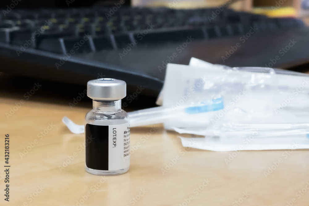 A bottle with Covid-19 vaccine stands on the table against the background of a disposable needle and a computer keyboard. A bottle with Covid-19 vaccine stands on the table against the background of a disposable needle and a computer keyboard.