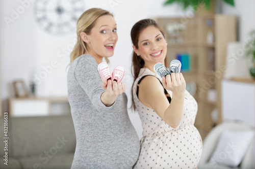two smiling pregnant women holding baby shoes
