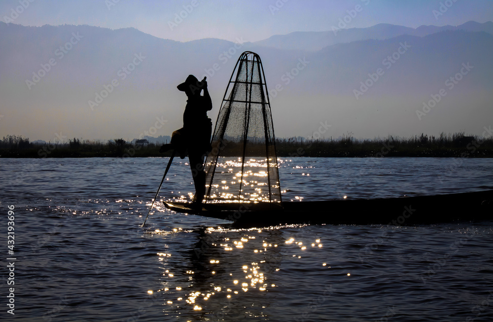 Silhouette of burmese traditional fisherman and basket standing on boat with paddle in the evening sun with glittering water reflections, blurred mountains background - Inle Lake, Myanmar