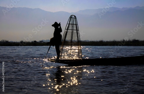 Silhouette of burmese traditional fisherman and basket standing on boat with paddle in the evening sun with glittering water reflections, blurred mountains background - Inle Lake, Myanmar