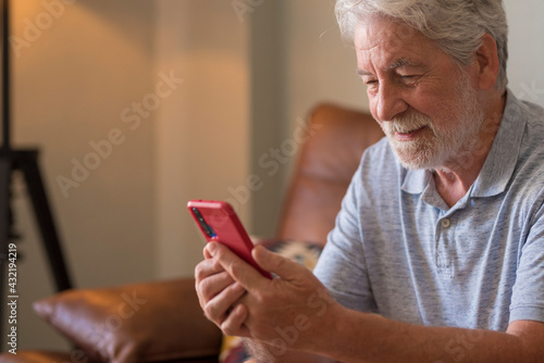 Happy senior handsome man in video call conference using modern smart phone sit down on the sofa at home - one mature male people with cellular connection technology lifestyle alone