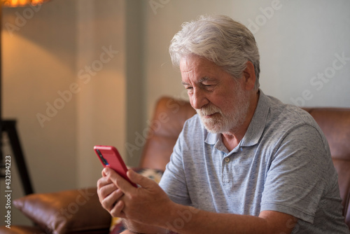 Mature handsome man text and write message on the smartphone - elderly people use cellular to chat sit down on the sofa at home - one retired people indoor lifestyle alone with connected device