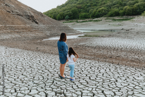 A woman and a girl stand, hand in hand, on dry, cracked ground and look at the lake, which is drying up. Global warming. © Oleksii Halutva