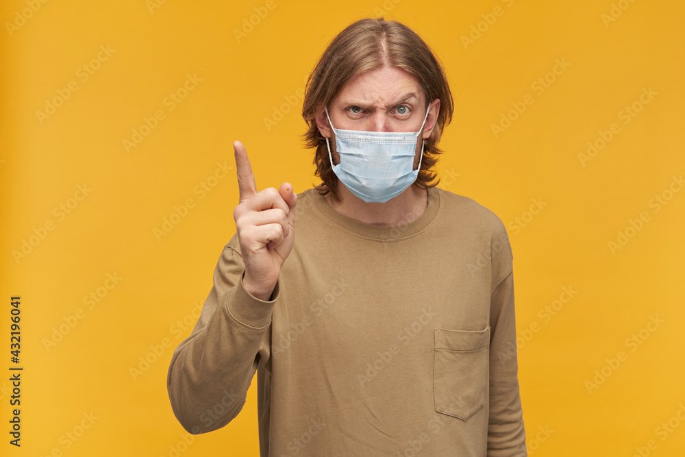 Unhappy guy with blond hair and beard. Wearing beige sweater and medical protective face mask. Threatens with a finger. Watching condemning at the camera isolated over yellow background Unhappy guy with blond hair and beard. Wearing beige sweater and medical protective face mask. Threatens with a finger. Watching condemning at the camera isolated over yellow background