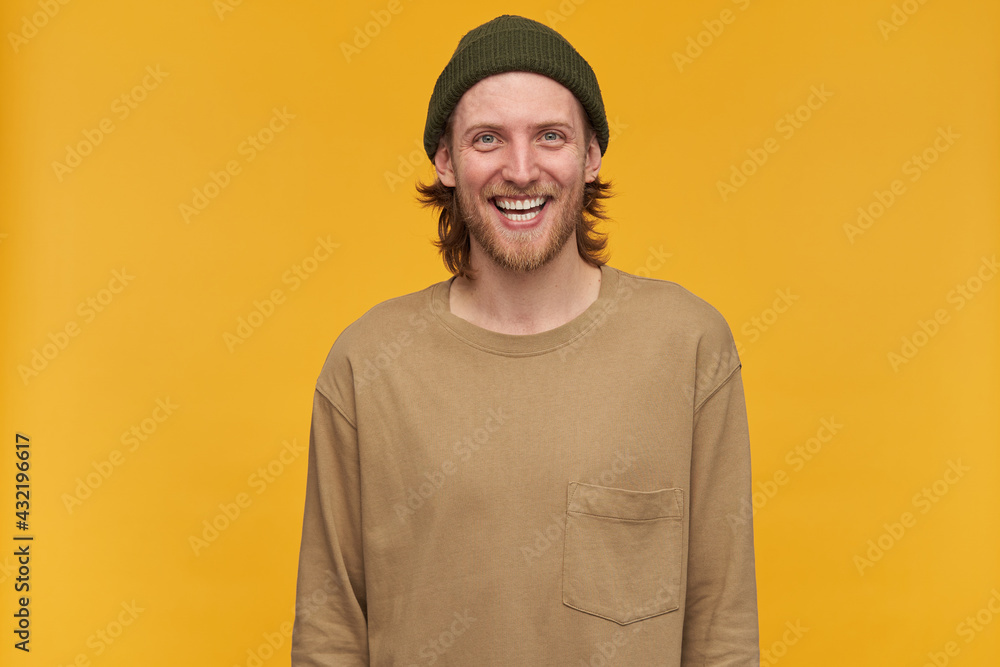 Young man, cheerful guy with blond hair, beard and mustache. Wearing green beanie and beige sweater. Watching at the camera and broadly smiling, isolated over yellow background Young man, cheerful guy with blond hair, beard and mustache. Wearing green beanie and beige sweater. Watching at the camera and broadly smiling, isolated over yellow background