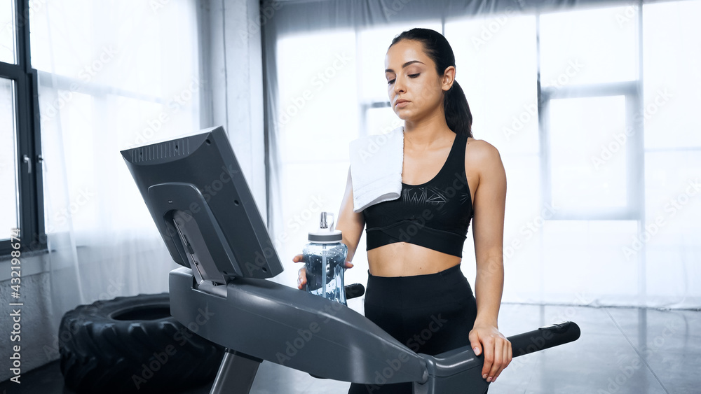 sweaty sportswoman with towel walking on treadmill and reaching sports bottle with water in gym. sweaty sportswoman with towel walking on treadmill and reaching sports bottle with water in gym.