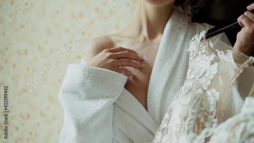 Caucasian woman in terry robe holds a wedding dress on a hanger in her hand. She gently strokes her bare shoulder in hotel room 