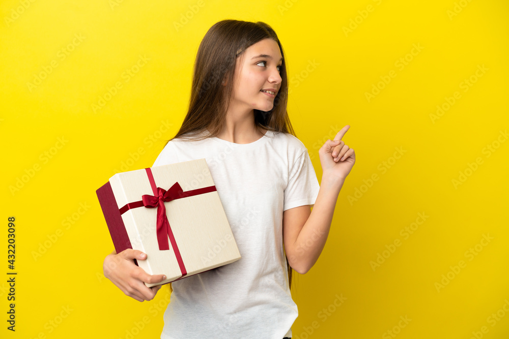 Little girl holding a gift over isolated yellow background pointing up a great idea Little girl holding a gift over isolated yellow background pointing up a great idea