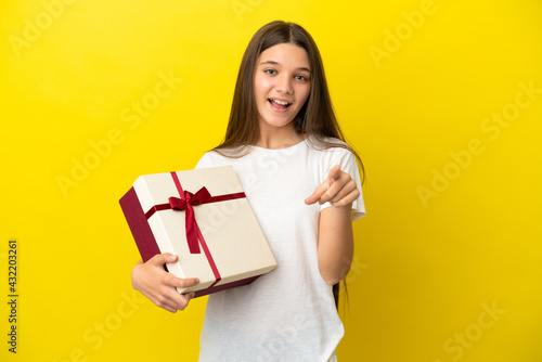 Little girl holding a gift over isolated yellow background surprised and pointing front