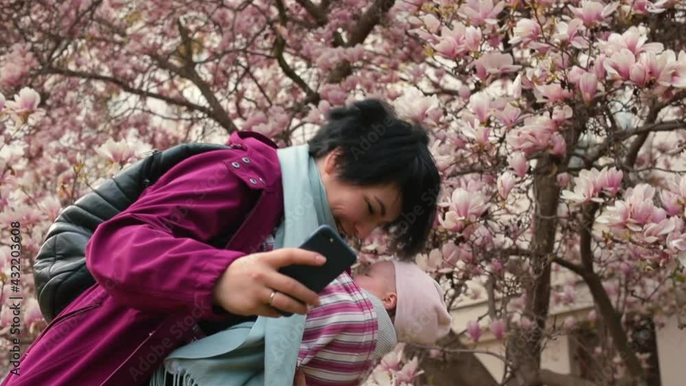 Mom in a sling holds the baby on her. She Takes pictures on the phone. Magnolia flowers in the background. Baby care. Mom in a sling holds the baby on her. She Takes pictures on the phone. Magnolia flowers in the background. Baby care.