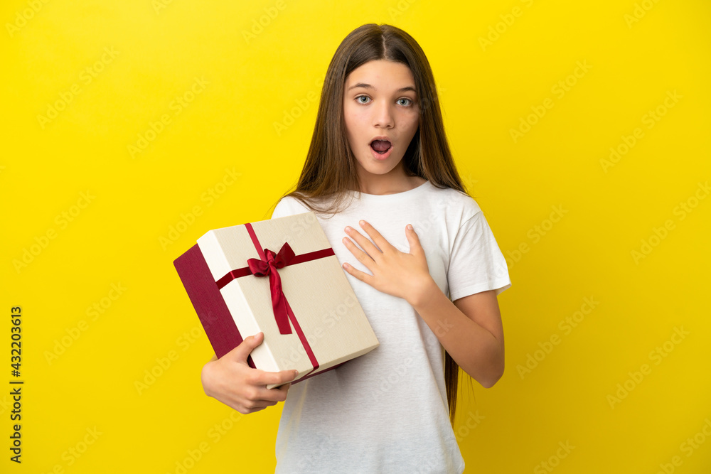 Little girl holding a gift over isolated yellow background surprised and shocked while looking right Little girl holding a gift over isolated yellow background surprised and shocked while looking right