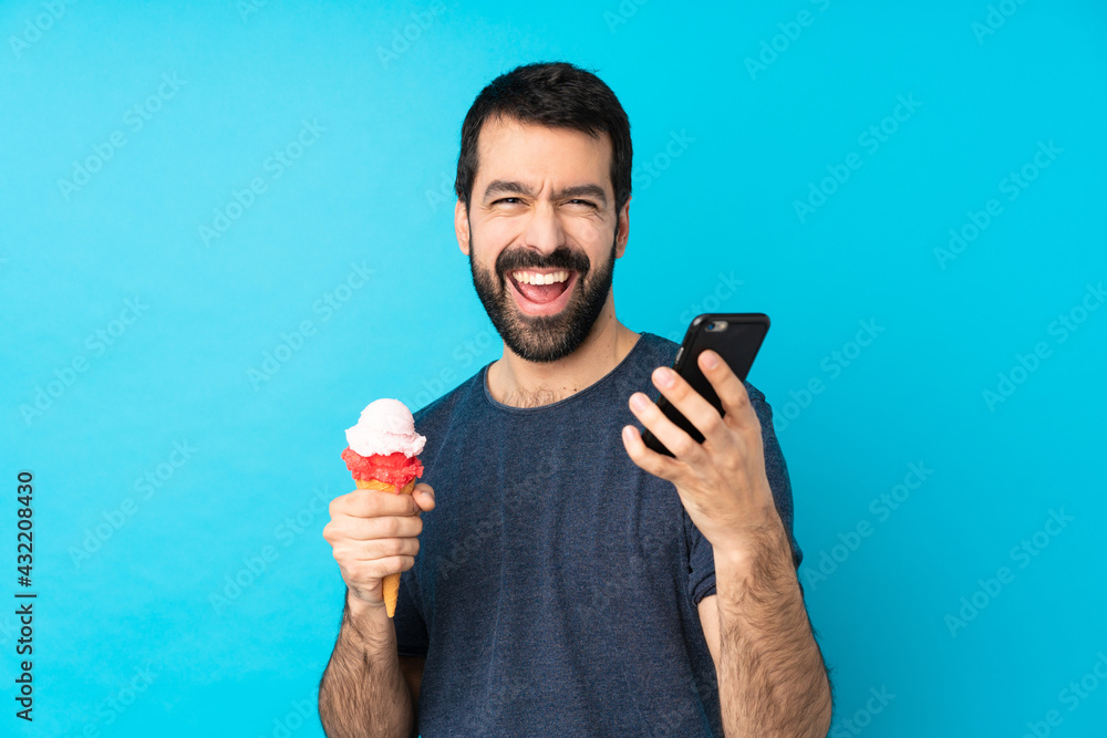 Young man with a cornet ice cream over isolated blue background with phone in victory position Young man with a cornet ice cream over isolated blue background with phone in victory position