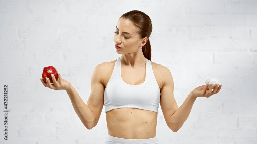 Young sportswoman with apple and tasty marshmallow near white wall. Young sportswoman with apple and tasty marshmallow near white wall.
