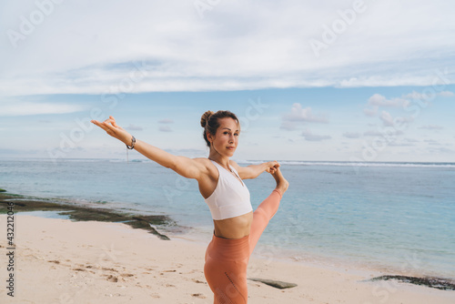 Flexible ethnic female performing Standing Big Toe Hold yoga asana on sandy coast