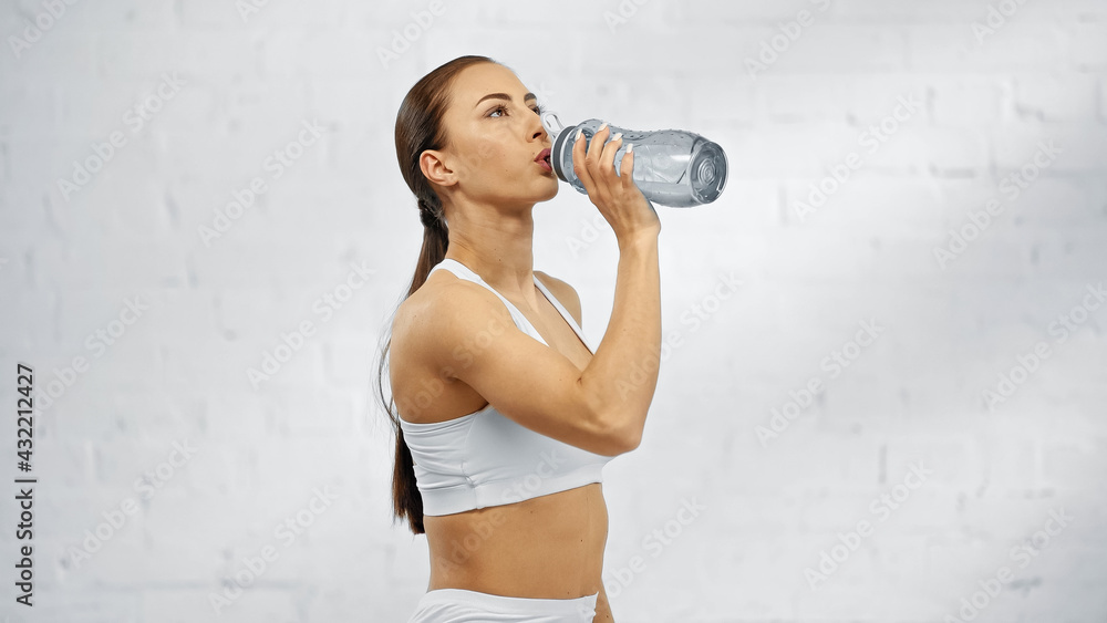 Sportswoman drinking water from sports bottle at home. - obrazy, fototapety, plakaty 