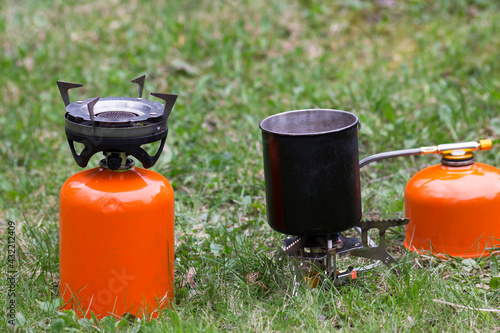Two camping gas stoves stand on the grass. There is a metal mug on one of the burners.