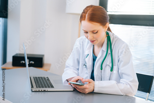 happy serious female doctor in white coat giving fast remote diagnose the patients using mobile phone, sitting at desk with laptop on background of window.