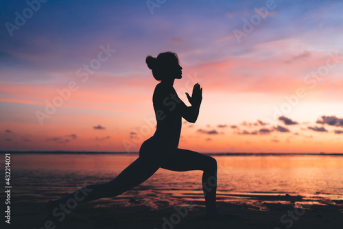 Side view of female silhouette keep praying during mindfulness meditation at seashore beach with colorful sky, slim woman in Namaste relaxing during evening physical recreation at coastline