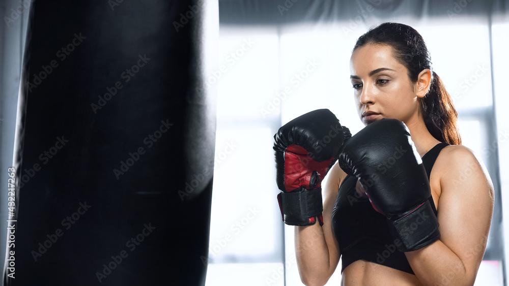 sportswoman in boxing gloves and sportswear exercising with punching bag in gym.