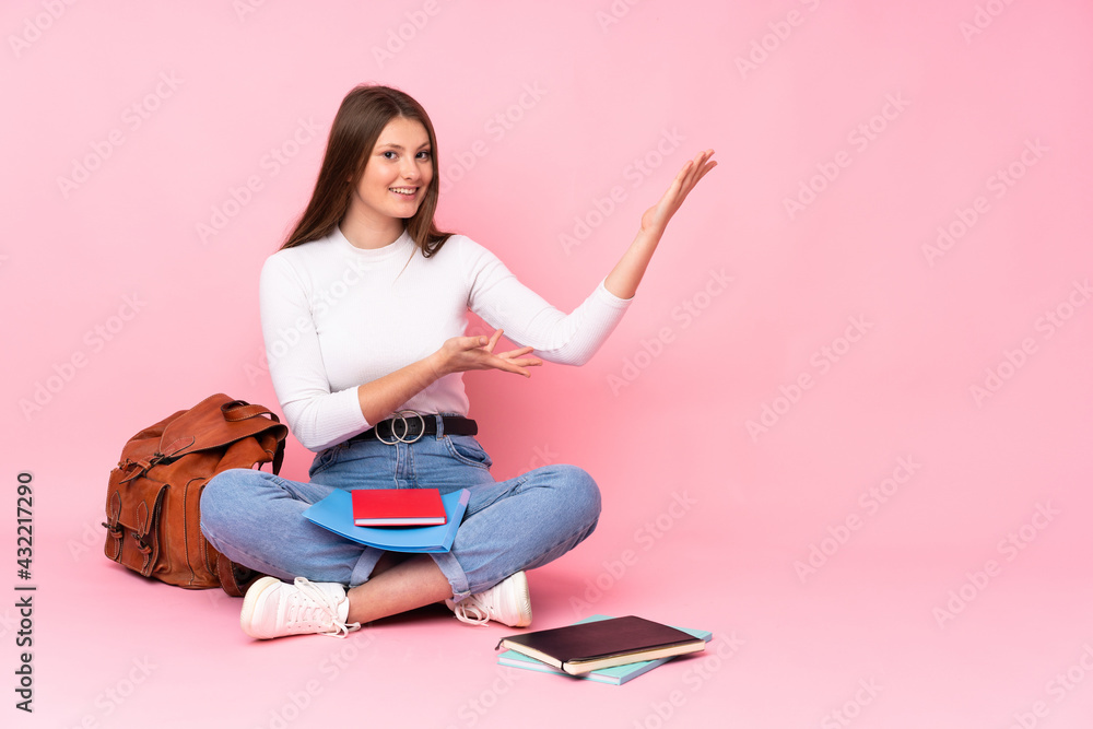 Teenager caucasian student girl sitting on the floor isolated on pink background extending hands to the side for inviting to come