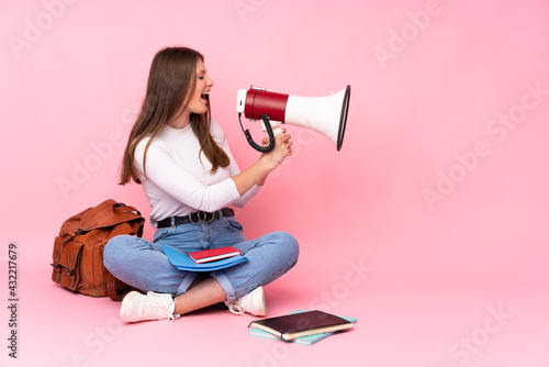 Teenager caucasian student girl sitting on the floor isolated on pink background shouting through a megaphone