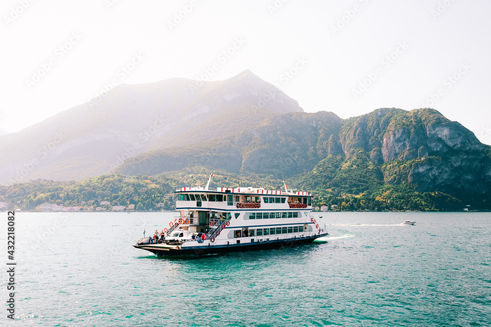 Peaceful Lake Como (Italian Lago di Como). Big ferry boat transportation with cars and people in amazing harbor. Small coastal resort town in Alps. Scenic panorama. Nautical vessel. Forest and hills.