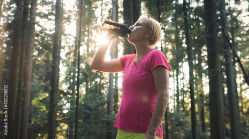 Middle shot of a Caucasian woman in sportswear standing in the forest, drinking water from a bottle