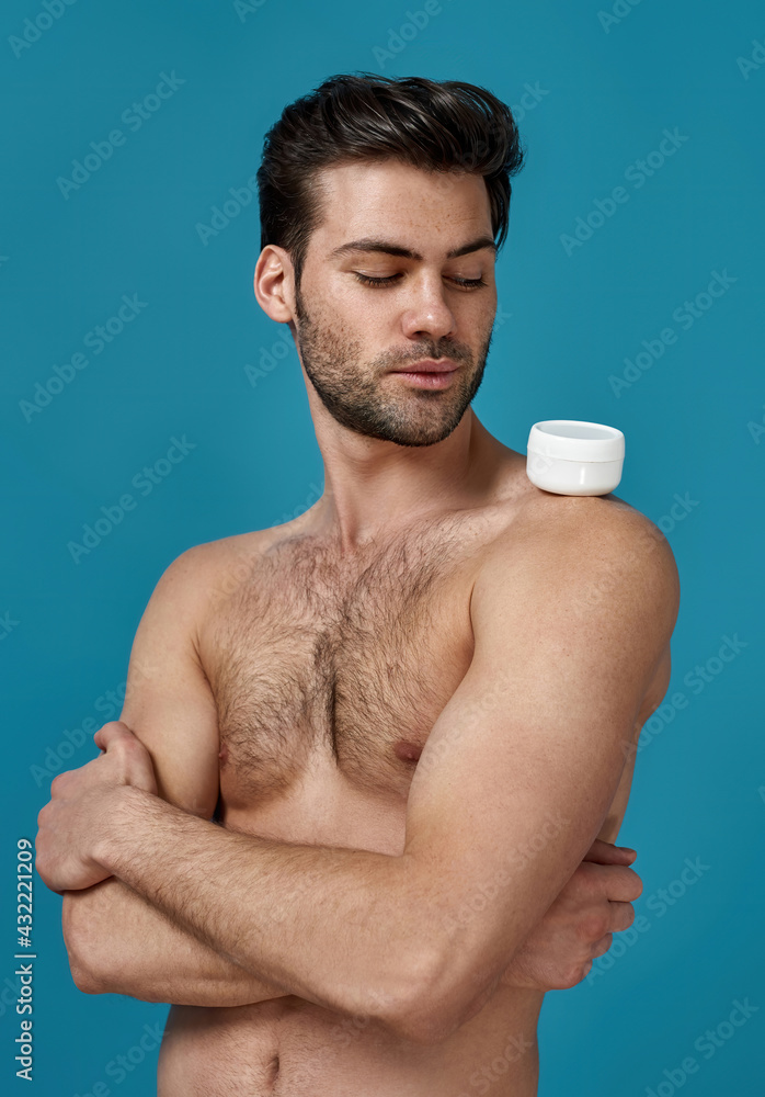 Fototapeta premium Studio shot of naked brunette guy looking at white jar of effective cream on his shoulder, posing with arms crossed isolated over blue background