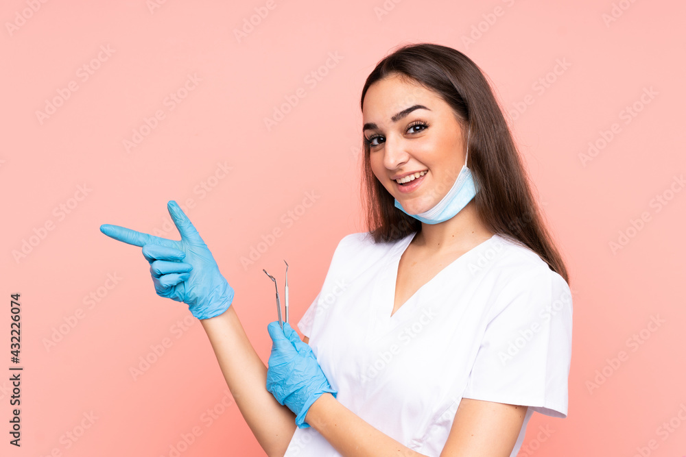 Woman dentist holding tools isolated on pink background pointing finger to the side Woman dentist holding tools isolated on pink background pointing finger to the side