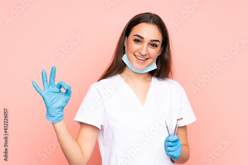 Woman dentist holding tools isolated on pink background showing an ok sign with fingers