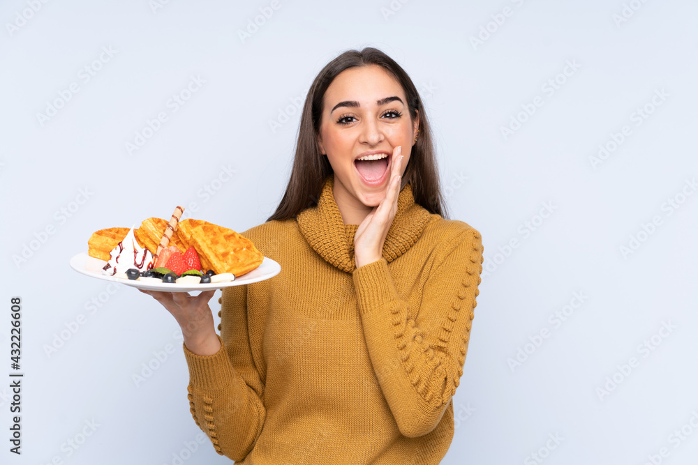 Young caucasian woman holding waffles isolated on blue background shouting with mouth wide open Young caucasian woman holding waffles isolated on blue background shouting with mouth wide open