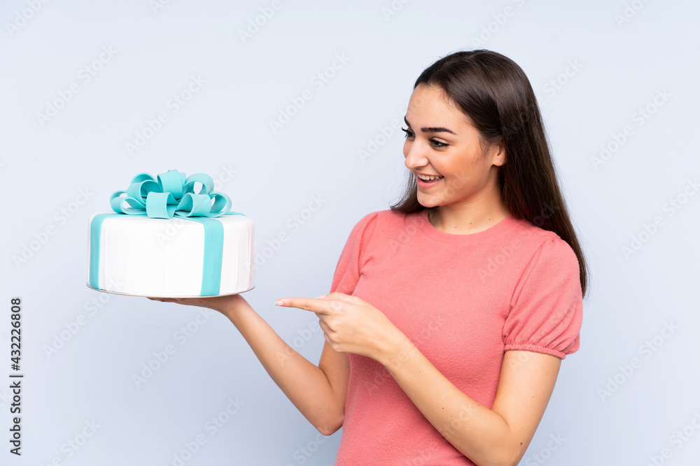 Pastry chef holding a big cake isolated on blue background pointing to the side to present a product Pastry chef holding a big cake isolated on blue background pointing to the side to present a product
