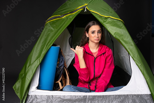 Young caucasian woman inside a camping green tent unhappy and pointing to the side