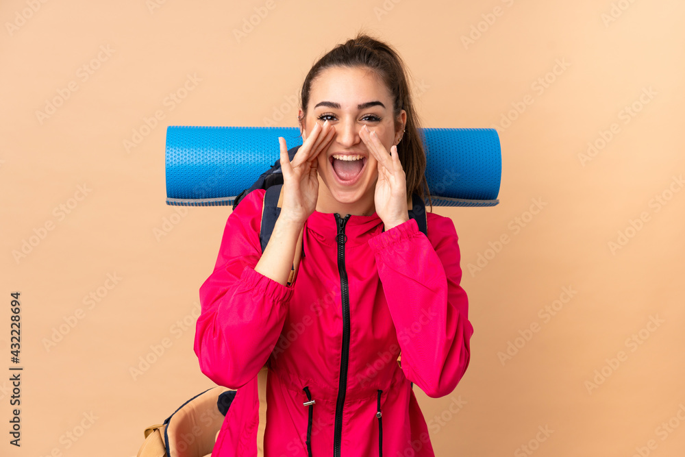 Young mountaineer girl with a big backpacker isolated on beige background shouting with mouth wide open Young mountaineer girl with a big backpacker isolated on beige background shouting with mouth wide open