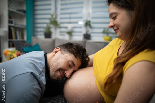 A husband puts his ear to his pregnant wife's belly to feel and hear their unborn child. A woman in advanced pregnancy. © fotodrobik